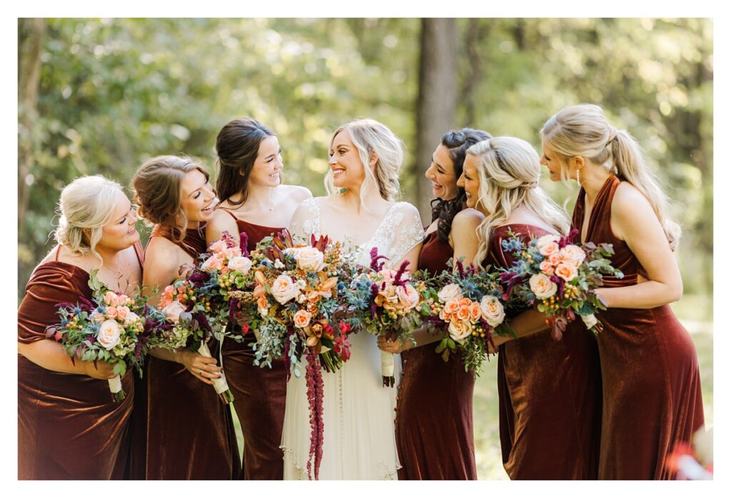 Group of bridesmaids in burgundy dresses with the bride, showcasing colorful fall flower bouquets at an outdoor wedding ceremony.